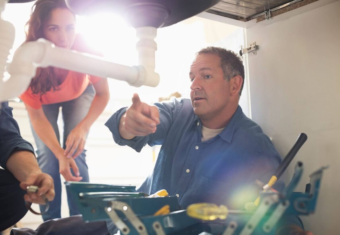 plumber pointing to a pipe under the kitchen sink so the female homeowner can see what the plumbing problem is